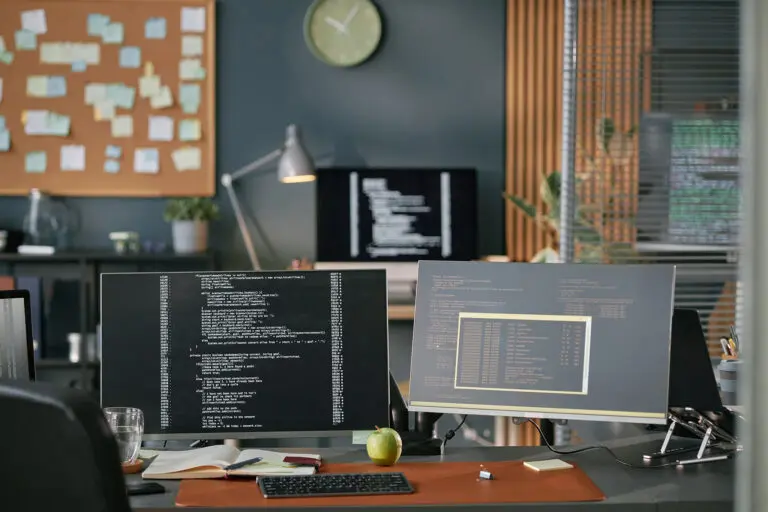 Modern office workspace featuring dual monitors displaying programming code, desktop computer, keyboard, notebook, green apple, glass of water, and blurred background with bulletin board